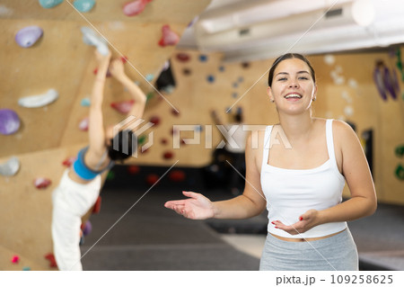 Positive slender europian girl in sportswear is standing near climbing wall in gym Positive slender europian girl in sportswear is standing near climbing wall in gym 109258625
