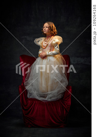 Portrait of little girl, kid dressed like aristocratic medieval person in old-fashioned dress stands on throne against dark vintage background. 109264168