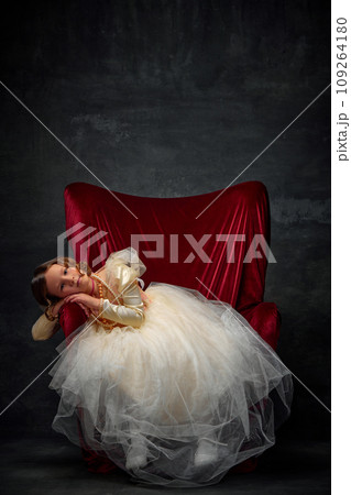 Cute little girl sitting in red throne dressed like medieval person in old fashioned dress and dreaming against dark vintage background. 109264180