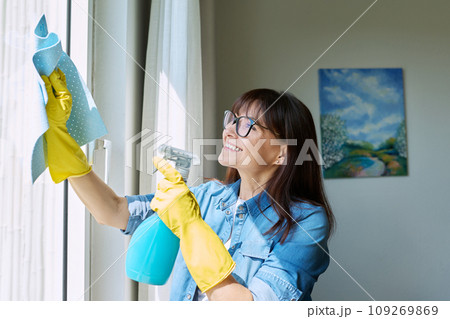 Middle-aged woman in gloves with spray cleaning windows at home Middle-aged woman in gloves with spray cleaning windows at home 109269869