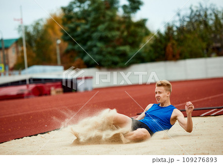 Athletic prowess on display: man mid-flight in long jump. Professional, skilled sportsman landing in sand court of impressive long jump Athletic prowess on display: man mid-flight in long jump. Professional, skilled sportsman landing in sand court of impressive long jump 109276893