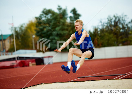 Graceful arch as a runner jumps into sand. Dynamic portrait of athletic man, professional sportsman doing perfect long jump. Graceful arch as a runner jumps into sand. Dynamic portrait of athletic man, professional sportsman doing perfect long jump. 109276894