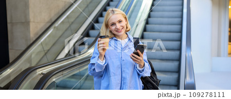 Image of beautiful female model, student standing near escalator, raising cup of takeaway coffee, smiling and looking happy, has mobile phone in hand and backpack on shoulder 109278111