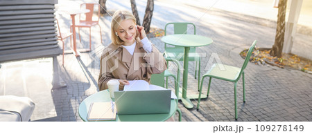 Lifestyle and technology concept. Young blond working woman in trench coat, studying, working on project on laptop, sitting in coffee shop on sunny day, making notes, attending online course 109278149