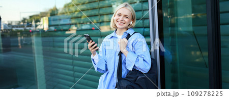 Lifestyle portrait of smiling young female model, student with backpack, waiting for someone on street, standing outdoors with smartphone Lifestyle portrait of smiling young female model, student with backpack, waiting for someone on street, standing outdoors with smartphone 109278225
