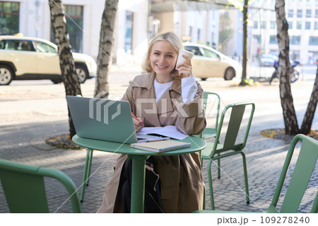 Portrait of young smiling blond woman, working on laptop, sitting in outdoor cafe on street, drinking coffee Portrait of young smiling blond woman, working on laptop, sitting in outdoor cafe on street, drinking coffee 109278240