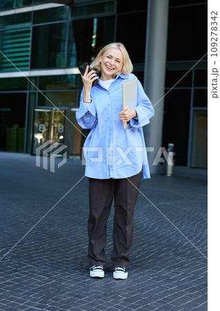 Vertical shot of modern, stylish young blond woman smiling, posing in trousers and blue collar shirt, holding smartphone and laughing, posing near campus building, has laptop in hand Vertical shot of modern, stylish young blond woman smiling, posing in trousers and blue collar shirt, holding smartphone and laughing, posing near campus building, has laptop in hand 109278342