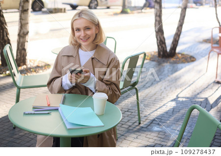 Image of young beautiful girl, student studying, doing homework in cafe, using mobile phone, holding smartphone, smiling Image of young beautiful girl, student studying, doing homework in cafe, using mobile phone, holding smartphone, smiling 109278437