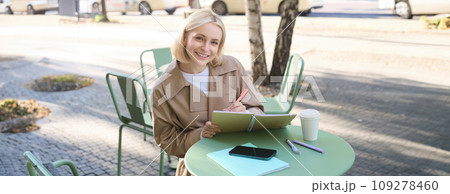 Portrait of beautiful blond woman, sitting in outdoor coffee shop, drawing in cafe in notebook, making sketches outside on street 109278460