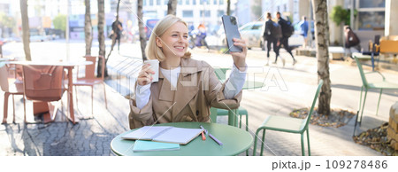 Happy blond woman taking selfie on smartphone, sitting outdoors in cafe and drinking coffee, making a post on social media to promote favourite shop Happy blond woman taking selfie on smartphone, sitting outdoors in cafe and drinking coffee, making a post on social media to promote favourite shop 109278486