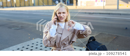 Disappointed young woman sitting on bench and showing thumbs down, talking on mobile phone and frowning, dislike conversation 109278541