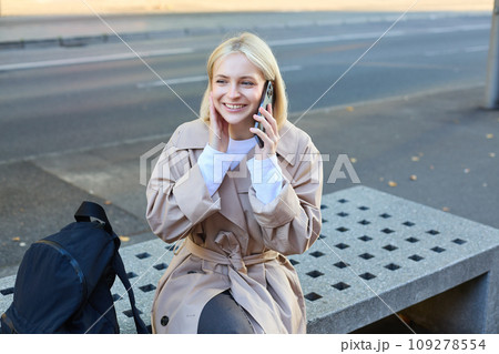 Image of young blond woman sitting on street bench with backpack, talking on mobile phone, answers a call and smiling while chatting with someone 109278554