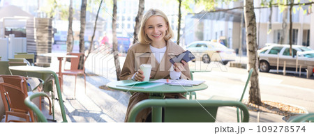 Portrait of modern young woman in city centre, sitting in cafe outdoors, drinking coffee, using mobile phone, smiling 109278574