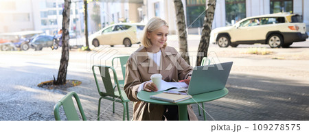 Image of young modern woman, sitting in cafe outdoors, drinking coffee or tea, looking at laptop, working on project, freelancing, making notes in notebook Image of young modern woman, sitting in cafe outdoors, drinking coffee or tea, looking at laptop, working on project, freelancing, making notes in notebook 109278575