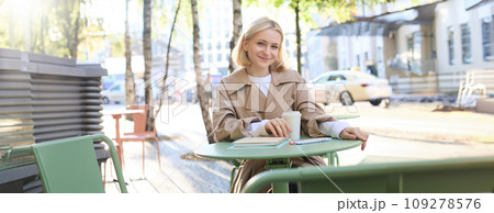 Portrait of young blonde woman sitting in cafe, doing homework and drinking coffee, writiing in journal, using notebook on fresh air Portrait of young blonde woman sitting in cafe, doing homework and drinking coffee, writiing in journal, using notebook on fresh air 109278576