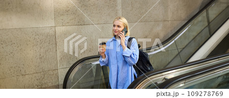 Young woman, student on escalator, drinking coffee and answer to phone call, talking with someone and smiling 109278769