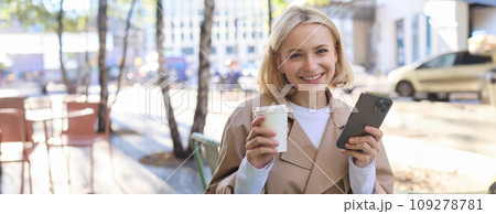 Portrait of cute blond woman with smartphone, drinking chai, coffee at favourite place in city centre, sitting outdoors, holding mobile phone 109278781