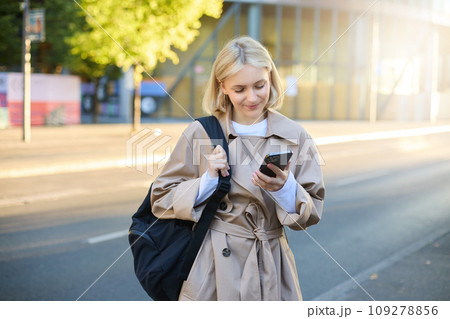 Image of young modern woman, student waiting for someone on street, standing with backpack and checking her mobile phone, looking at smartphone screen Image of young modern woman, student waiting for someone on street, standing with backpack and checking her mobile phone, looking at smartphone screen 109278856