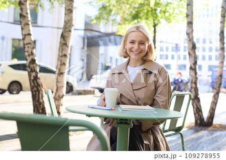 Urban lifestyle concept. Young beautiful woman sitting in outdoor cafe, contemplating city life, drinking coffee and smiling on street Urban lifestyle concept. Young beautiful woman sitting in outdoor cafe, contemplating city life, drinking coffee and smiling on street 109278955