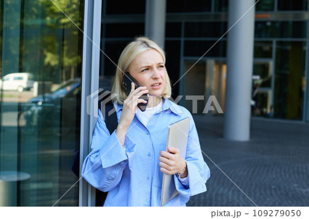 Portrait of young modern woman, office manager near building, standing outside with backpack, laptop, talking on mobile phone, chatting on smartphone with confused face 109279050