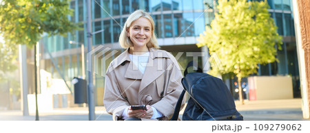 Lifestyle portrait of smiling female model sitting on street bench with backpack and mobile phone, waiting for friend and scrolling social media on smartphone 109279062