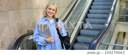 Image of young modern woman, student on escalator, holding laptop and backpack, walking in city 109279063