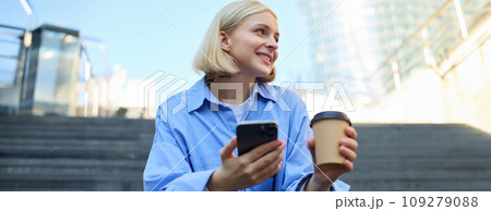 Lifestyle picture of smiling, beautiful young woman, drinking coffee, using mobile phone, resting in city, sitting on stairs 109279088