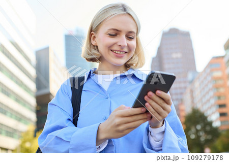 Close up portrait of stylish young blonde woman, standing on street, checking her mobile phone, using smartphone app to get around town, looking at online map for directions 109279178