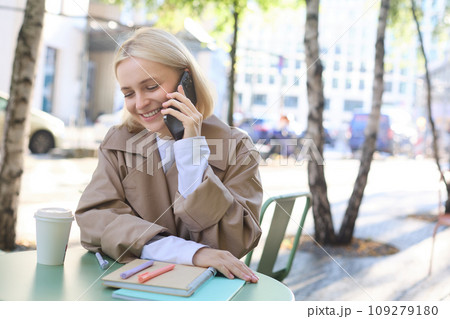 Portrait of young beautiful woman in cafe, sitting outdoors on bright sunny day, talking on smartphone, chatting over the phone and smiling 109279180
