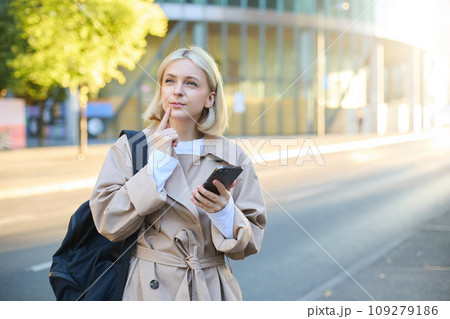 Portrait of woman with thinking face, standing with backpack on street, holding smartphone, pondering, making a decision with thoughtful face Portrait of woman with thinking face, standing with backpack on street, holding smartphone, pondering, making a decision with thoughtful face 109279186