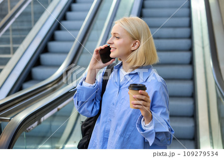 Portrait of cute young blond woman, student with cup of coffee, answers phone call, standing near escalator in city and talking, chatting with someone over the smartphone Portrait of cute young blond woman, student with cup of coffee, answers phone call, standing near escalator in city and talking, chatting with someone over the smartphone 109279534