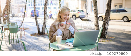Portrait of young happy woman, looking pleased and surprised at laptop screen, sitting in outdoor cafe, street coffee shop, holding hands on chest and smiling grateful 109279600