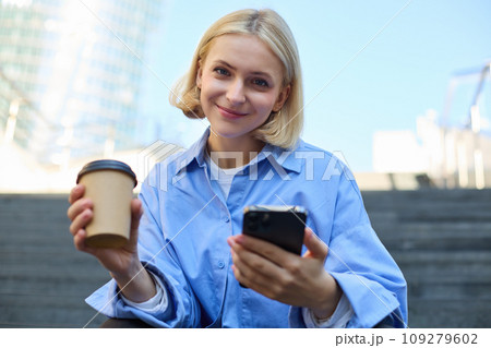 Portrait of beautiful blond woman, smiling girl student drinking coffee, sitting on stairs with smartphone, looking happy at camera 109279602