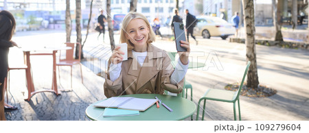 Lifestyle shot of stylish blond girl with smartphone, drinking coffee, posing with her chai for social media, creating content on phone, taking selfie 109279604