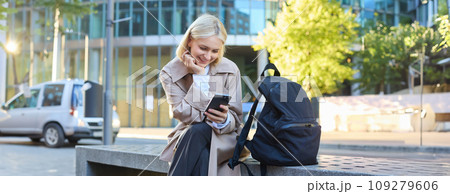 Lifestyle portrait of cute blonde girl with smartphone, sitting on bench with backpack, using mobile phone, reading message 109279606
