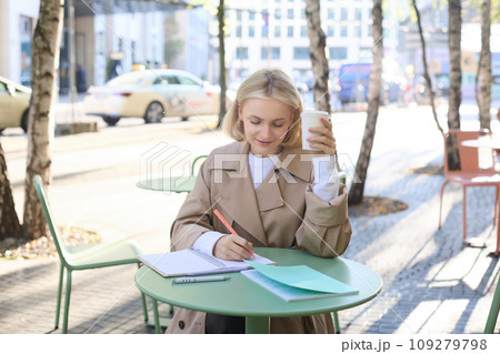 Portrait of young professional, female student working on project outdoors, doing homework, drinking coffee and sitting in cafe outside on street Portrait of young professional, female student working on project outdoors, doing homework, drinking coffee and sitting in cafe outside on street 109279798