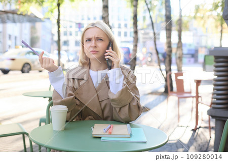 Portrait of woman, student doing homework outdoors in cafe, holding pen, frowning while calling someone, struggling to understand phone conversation Portrait of woman, student doing homework outdoors in cafe, holding pen, frowning while calling someone, struggling to understand phone conversation 109280014