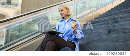Image of stylish young modern woman, student doing homework, studying outdoors on campus stairs, sitting with laptop and coffee, drinking her cappuccino and connecting to public wifi 109280053