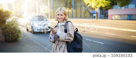 Stylish young blonde woman, girl with smartphone and backpack, standing on street, using mobile phone application, posing close to the road with cars and sky scrappers Stylish young blonde woman, girl with smartphone and backpack, standing on street, using mobile phone application, posing close to the road with cars and sky scrappers 109280102