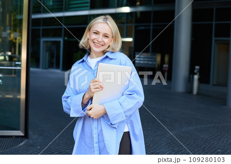Image of young female employee, working woman standing near her office on street, holding pile of documents and smiling Image of young female employee, working woman standing near her office on street, holding pile of documents and smiling 109280103