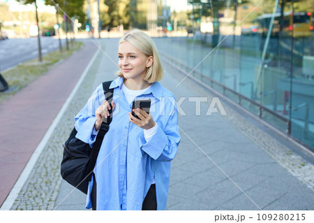 Portrait of blond young woman, using navigation map app on smartphone, walking along street, using directions on phone, holding backpack 109280215