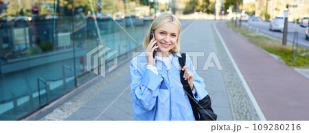 Portrait of young smiling woman talking on mobile phone, student walks home with smartphone, carries black backpack 109280216