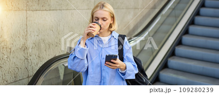 A happy, elegant businesswoman descending the escalator and holding takeaway coffee while reading important messages. A woman on the escalator with a phone 109280239