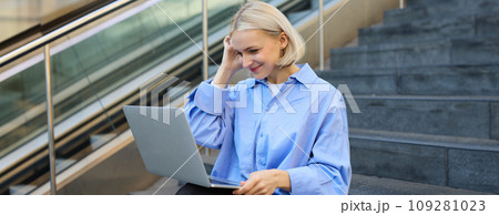 Young woman, student in blue shirt, working on laptop, sitting outdoors on street stairs, working on project online, connects to public wifi, elearning Young woman, student in blue shirt, working on laptop, sitting outdoors on street stairs, working on project online, connects to public wifi, elearning 109281023