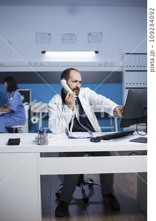 In medical office, male physician is using a computer for medical research and chatting on a wired phone. Caucasian man in lab coat using landline phone to speak with hospital receptionist. 109284092