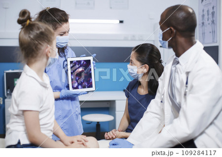 In the clinic, female nurse grasps a digital tablet showing a bacteria image in front of the patients and African American doctor. Expert care and education ensure healthcare and wellness. 109284117