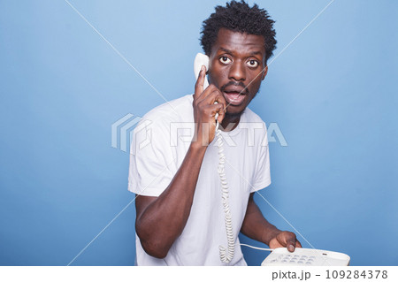 Surprised young black man with afro hair speaks on a telephone in a studio. African american guy holds landline phone in hand against blue background, expressing shock during conversation. 109284378