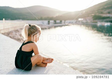 Little barefoot girl sits cross-legged on the pier and looks at the water. Side view Little barefoot girl sits cross-legged on the pier and looks at the water. Side view 109284781