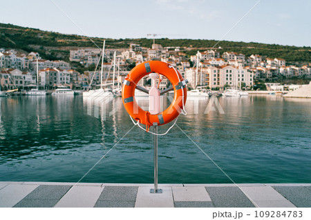 Orange lifebuoy on a stand on a pier against the backdrop of a colorful resort town with moored yachts Orange lifebuoy on a stand on a pier against the backdrop of a colorful resort town with moored yachts 109284783