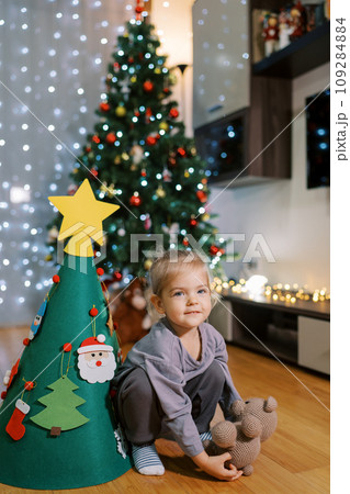 Little girl squats with a teddy bear near a decorated toy Christmas tree Little girl squats with a teddy bear near a decorated toy Christmas tree 109284884